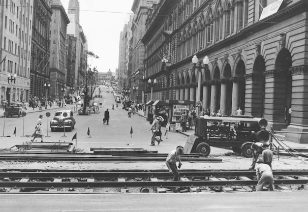 A busy urban street is lined with tall, ornate buildings on both sides. Several pedestrians walk along the sidewalks and cross the street, while a few cars are visible driving down the road. In the foreground, construction workers are actively working on railroad tracks, with some machinery nearby.