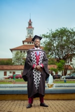 A person in a graduation gown and cap stands confidently in front of a clock tower. The background includes a fountain, trees, and parked cars, creating an academic setting.