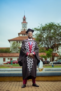 A person in a graduation gown and cap stands confidently in front of a clock tower. The background includes a fountain, trees, and parked cars, creating an academic setting.