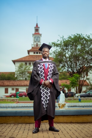 A person in a graduation gown and cap stands confidently in front of a clock tower. The background includes a fountain, trees, and parked cars, creating an academic setting.