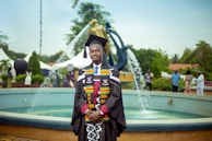 A graduate in academic regalia stands in front of a fountain. The regalia includes a black cap and gown, with colorful stoles. The background features a modern sculpture and several people casually dressed in the vicinity, suggesting an outdoor event, likely a graduation ceremony.