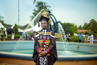 A graduate in academic regalia stands in front of a fountain. The regalia includes a black cap and gown, with colorful stoles. The background features a modern sculpture and several people casually dressed in the vicinity, suggesting an outdoor event, likely a graduation ceremony.