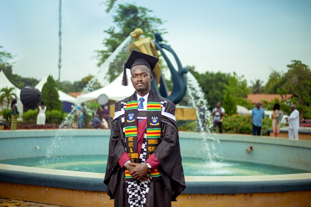 A graduate in academic regalia stands in front of a fountain. The regalia includes a black cap and gown, with colorful stoles. The background features a modern sculpture and several people casually dressed in the vicinity, suggesting an outdoor event, likely a graduation ceremony.