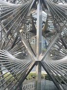 Intricate metal sculpture with geometric design elements is situated in an urban setting. The metallic structure features a series of intersecting lines and angles, creating a complex, abstract form. In the background, modern buildings and greenery are visible, providing contrast to the industrial texture of the sculpture.