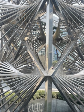 Intricate metal sculpture with geometric design elements is situated in an urban setting. The metallic structure features a series of intersecting lines and angles, creating a complex, abstract form. In the background, modern buildings and greenery are visible, providing contrast to the industrial texture of the sculpture.
