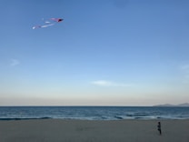 A beach scene depicting a child flying a red kite against a clear blue sky. The ocean is calm, and the scene is peaceful with distant mountains visible on the horizon. The child is standing on the sandy shore, focusing on the kite.