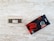 A 1TB SSD module inside a clear plastic case is placed on a textured light wooden surface. Next to it is the product's black and red packaging box, labeled with specifications and featuring a stylized logo.