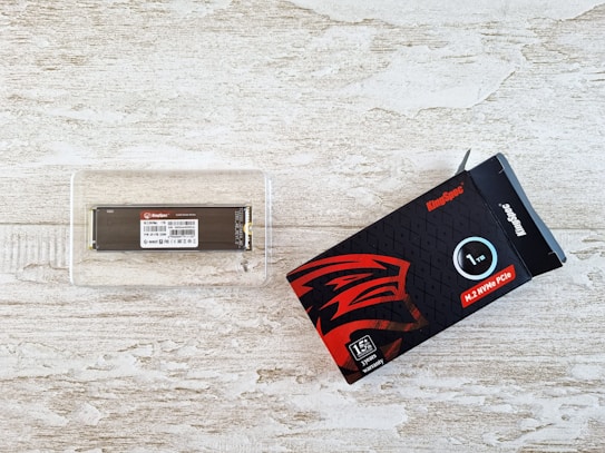 A 1TB SSD module inside a clear plastic case is placed on a textured light wooden surface. Next to it is the product's black and red packaging box, labeled with specifications and featuring a stylized logo.