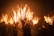 A group of people holds large torches made of multiple wooden sticks burning brightly. The dark background emphasizes the fiery glow and smoke, while their faces are partly visible under helmets or hats.