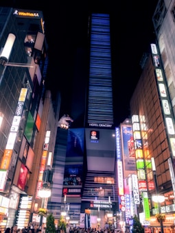 A bustling city scene at night featuring a tall building with a prominent 'Hotel Gracery' sign and an eye-catching Godzilla head emerging from the top. Surrounding the building are other high-rise structures, each adorned with vibrant, colorful neon advertisements in various languages. Below, a busy street is filled with people and illuminated by streetlights and shop signs.