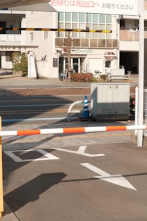A street scene with a parking or security barrier in the foreground. The barrier is red and white, and there are nearby traffic markings on the road. In the background, there is a building with multiple windows and a sign in Japanese script. A blue traffic cone is also present next to some shrubs and flowers.
