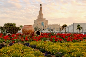 A landscaped garden featuring vibrant red and yellow flowers in the foreground with large clay pots adding rustic decoration. The backdrop includes a distinctive spiral mosque structure, palm trees, and a clear sky with scattered clouds.