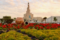 A landscaped garden featuring vibrant red and yellow flowers in the foreground with large clay pots adding rustic decoration. The backdrop includes a distinctive spiral mosque structure, palm trees, and a clear sky with scattered clouds.