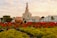 A landscaped garden featuring vibrant red and yellow flowers in the foreground with large clay pots adding rustic decoration. The backdrop includes a distinctive spiral mosque structure, palm trees, and a clear sky with scattered clouds.