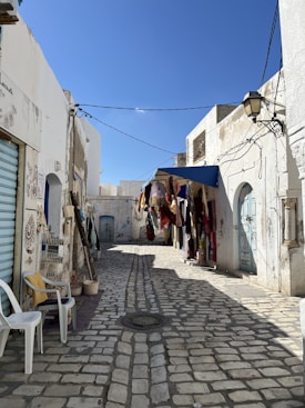 A narrow cobblestone street in a traditional, sunlit neighborhood. The walls of the buildings are painted white, with some having colorful doors in shades of blue. Laundry or textiles hang from a line strung across the street, adding vibrant colors to the scene. Two plastic chairs are placed against a wall on the left side. A vintage street lamp is affixed to a building on the right, and overhead wires crisscross the alleyway.