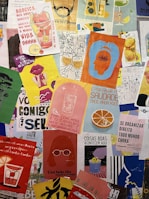 A collection of colorful posters and artwork covers a surface, featuring various illustrations, text in Portuguese, and vibrant designs. The posters include images of drinks, abstract shapes, human figures, and a mix of colors and styles, creating a lively and artistic collage.