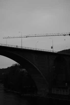A large bridge with an arched under-structure spans across a river, silhouetted against a cloudy sky. A construction crane is positioned to the right, parallel to the bridge. Street lamps and a pedestrian are visible on the bridge.