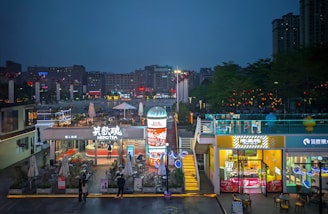 A vibrant urban area illuminated by various signs and lights, featuring a busy commercial space with businesses like a tea shop called Hero Tea. People are seen engaging in activities, and there are several colorful billboards and advertisements. The scene is set during the evening, with the sky transitioning to night.