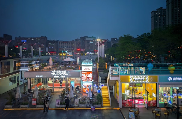 A vibrant urban area illuminated by various signs and lights, featuring a busy commercial space with businesses like a tea shop called Hero Tea. People are seen engaging in activities, and there are several colorful billboards and advertisements. The scene is set during the evening, with the sky transitioning to night.