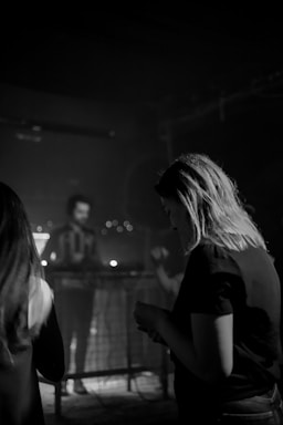A black and white scene depicting people in a dimly lit indoor space, possibly a club or concert venue. One person in the foreground is facing downward, with blurred figures and a musician or DJ in the background. The atmosphere is somewhat somber and introspective.