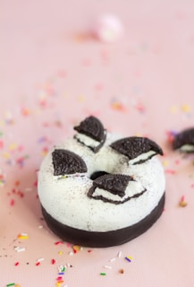 A chocolate donut with white icing and topped with pieces of chocolate cookies is placed on a light pink surface. Colorful sprinkles are scattered around the donut, adding a playful element to the scene.