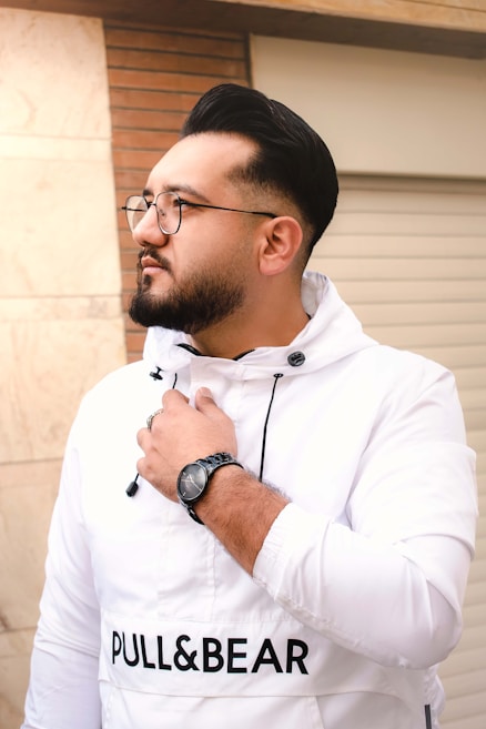 A person with short dark hair and a beard, wearing glasses and a white jacket with the brand name 'Pull&Bear' printed on it. The individual is wearing a black watch and appears to be outdoors, standing near a brick wall and a light-colored surface.