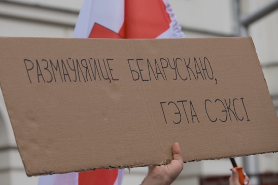 A hand is holding a cardboard sign with text written in Belarusian. In the background, part of a red and white flag is visible.