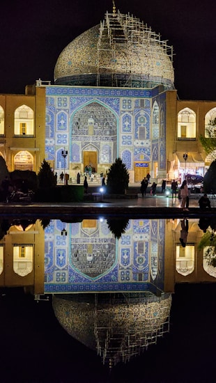 A beautifully illuminated mosque with intricate blue tile work and a large dome under restoration, reflected in a calm body of water below. People are visible near the entrance, while soft lighting highlights the architectural details at night.