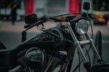 A close-up view of a black motorcycle with shiny chrome elements, focusing on the handlebars and the headlight. The brand name can be seen faintly on the fuel tank, and the background is slightly blurred, indicating a street scene.