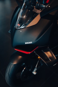 A close-up view of the front section of a Ducati motorcycle. The image focuses on the sleek, matte black fairing with a clear windshield, displaying the Ducati logo prominently. The design includes sharp angles and a touch of red accent underneath the headlight area. The tire and part of the wheel are also visible.