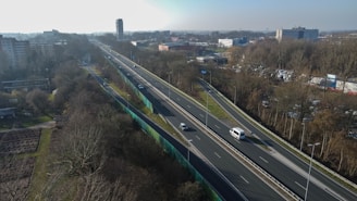 Aerial view of a highway cutting through a wooded area with several buildings visible in the background. Several vehicles are traveling along the highway, which is flanked by trees and a sound barrier on one side. The scene includes urban infrastructure with residential and commercial buildings.