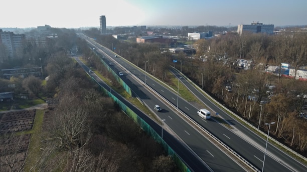 Aerial view of a highway cutting through a wooded area with several buildings visible in the background. Several vehicles are traveling along the highway, which is flanked by trees and a sound barrier on one side. The scene includes urban infrastructure with residential and commercial buildings.