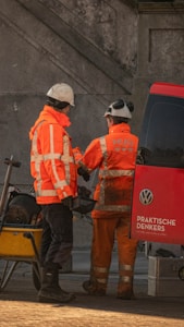 Two construction workers in bright orange uniforms and hard hats stand beside a red van with a 'Volkswagen' logo. They seem to be interacting, possibly reviewing plans or tools. A wheelbarrow filled with equipment is parked nearby on a cobblestone surface. The background features a concrete wall.