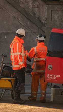 Two construction workers in bright orange uniforms and hard hats stand beside a red van with a 'Volkswagen' logo. They seem to be interacting, possibly reviewing plans or tools. A wheelbarrow filled with equipment is parked nearby on a cobblestone surface. The background features a concrete wall.