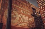 A vintage advertisement painted on a weathered brick wall, displaying a faded logo and the words 'Drink Miller'. The wall features a fire escape with metal railings, casting shadows across the brick surface. The signage reflects an old-time aesthetic, with muted colors and an aged appearance.