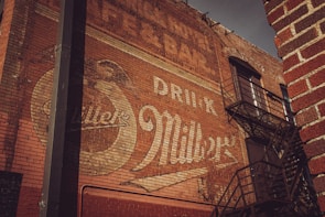 A vintage advertisement painted on a weathered brick wall, displaying a faded logo and the words 'Drink Miller'. The wall features a fire escape with metal railings, casting shadows across the brick surface. The signage reflects an old-time aesthetic, with muted colors and an aged appearance.