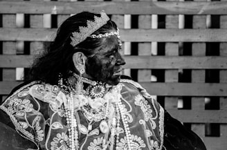 A person wearing elaborate traditional attire, including a crown, earrings, and a necklace, gazes to the side. The clothing features intricate patterns, and the image is in black and white. The background consists of a wooden lattice.
