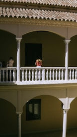 An architectural scene featuring a building with a tiled roof and arched openings. The image depicts a two-story structure with columns and white balustrades. A person stands on the upper level, wearing a red outfit, while the lower level contains decorative elements around a window.
