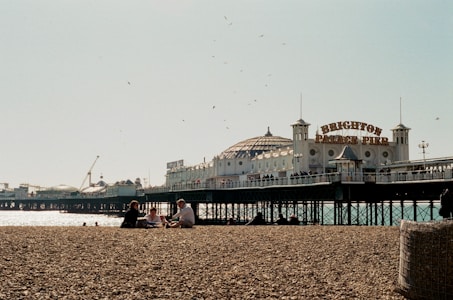 A pebble beach with several people sitting and relaxing, in the background is a pier extending over the water. The sign 'Brighton Palace Pier' is prominently displayed on the structure, with multiple people visible walking along the pier. Gulls are flying in the sky above, and the ocean sparkles under the sunlight.