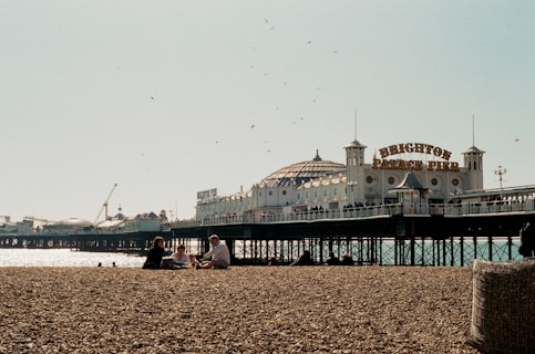 A pebble beach with several people sitting and relaxing, in the background is a pier extending over the water. The sign 'Brighton Palace Pier' is prominently displayed on the structure, with multiple people visible walking along the pier. Gulls are flying in the sky above, and the ocean sparkles under the sunlight.