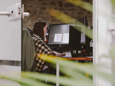 A woman working calmly at her desk with a notebook and laptop, surrounded by plants.