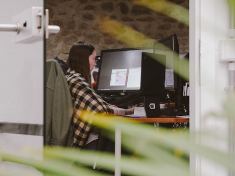 A person is seated at a desk in front of multiple computer monitors, working attentively. The workspace is framed by greenery from a plant in the foreground, and the surrounding environment features a stone wall, adding a rustic touch.