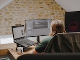 A person with long hair sits at a desk facing two computer screens. The workspace is set in a room with a rustic stone wall. Open laptops and programming code are visible on the screens. A white mug with a black pattern and some small objects are on the desk.