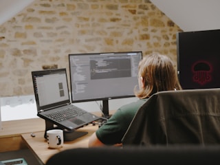 A person with long hair sits at a desk facing two computer screens. The workspace is set in a room with a rustic stone wall. Open laptops and programming code are visible on the screens. A white mug with a black pattern and some small objects are on the desk.