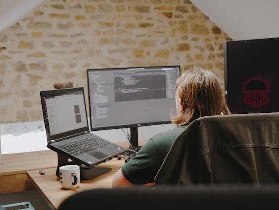 A person with long hair sits at a desk facing two computer screens. The workspace is set in a room with a rustic stone wall. Open laptops and programming code are visible on the screens. A white mug with a black pattern and some small objects are on the desk.