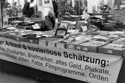 A black and white image of a market stall filled with various items such as books, postcards, stamps, and old currency. The items are displayed in boxes and on the table, with a large banner advertising their purchase and free appraisal. People are seen in the background, creating a busy atmosphere.