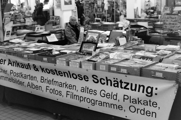 A black and white image of a market stall filled with various items such as books, postcards, stamps, and old currency. The items are displayed in boxes and on the table, with a large banner advertising their purchase and free appraisal. People are seen in the background, creating a busy atmosphere.