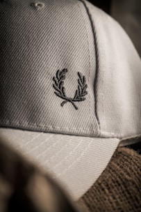 A close-up of a textured fabric cap featuring an embroidered laurel wreath design. The lighting creates a soft shadow effect, highlighting the intricate stitching and the cap's material.