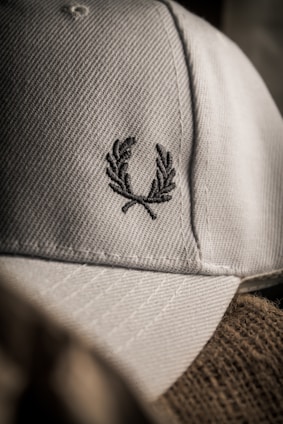 A close-up of a textured fabric cap featuring an embroidered laurel wreath design. The lighting creates a soft shadow effect, highlighting the intricate stitching and the cap's material.