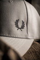 A close-up of a textured fabric cap featuring an embroidered laurel wreath design. The lighting creates a soft shadow effect, highlighting the intricate stitching and the cap's material.